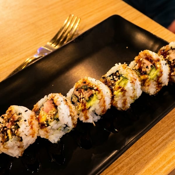 A black rectangular plate holds eight sushi rolls drizzled with sauce and sesame seeds.