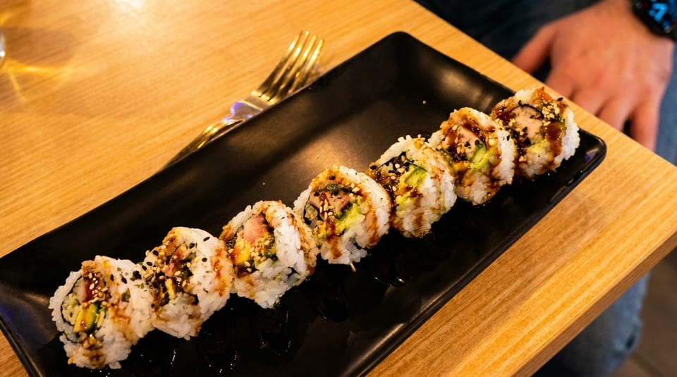 A black rectangular plate holds eight sushi rolls drizzled with sauce and sesame seeds.
