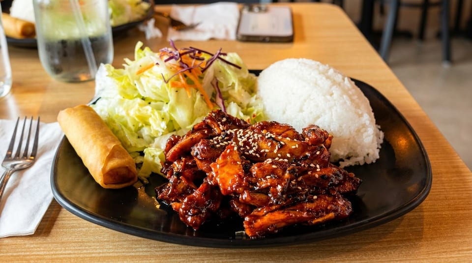 Teriyaki chicken with white rice, a side salad, and an egg roll on a black plate.