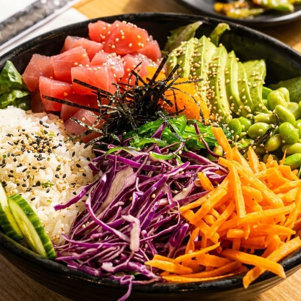 A colorful poke bowl with tuna, avocado, edamame, carrots, cabbage, and cucumbers over rice.