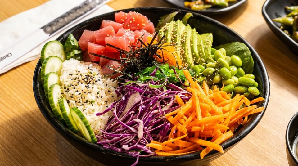 A colorful poke bowl with tuna, avocado, edamame, carrots, cabbage, and cucumbers over rice.
