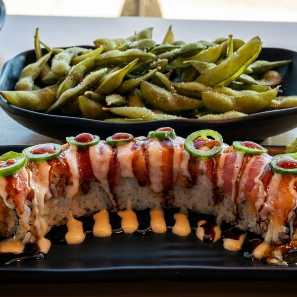 Sushi roll topped with fish, sauce, and jalapeños, with a side of edamame.