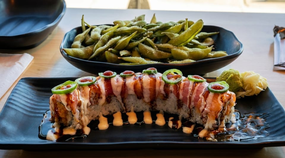 Sushi roll topped with fish, sauce, and jalapeños, with a side of edamame.