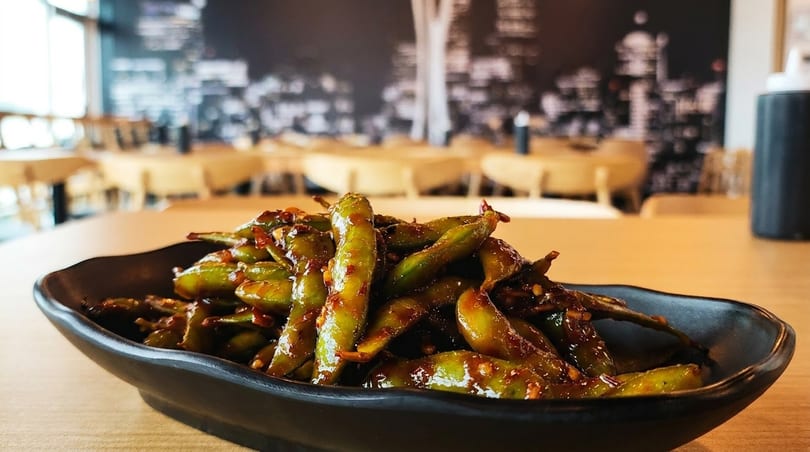 Spicy edamame in a black bowl on a wooden table with a restaurant interior in the background.