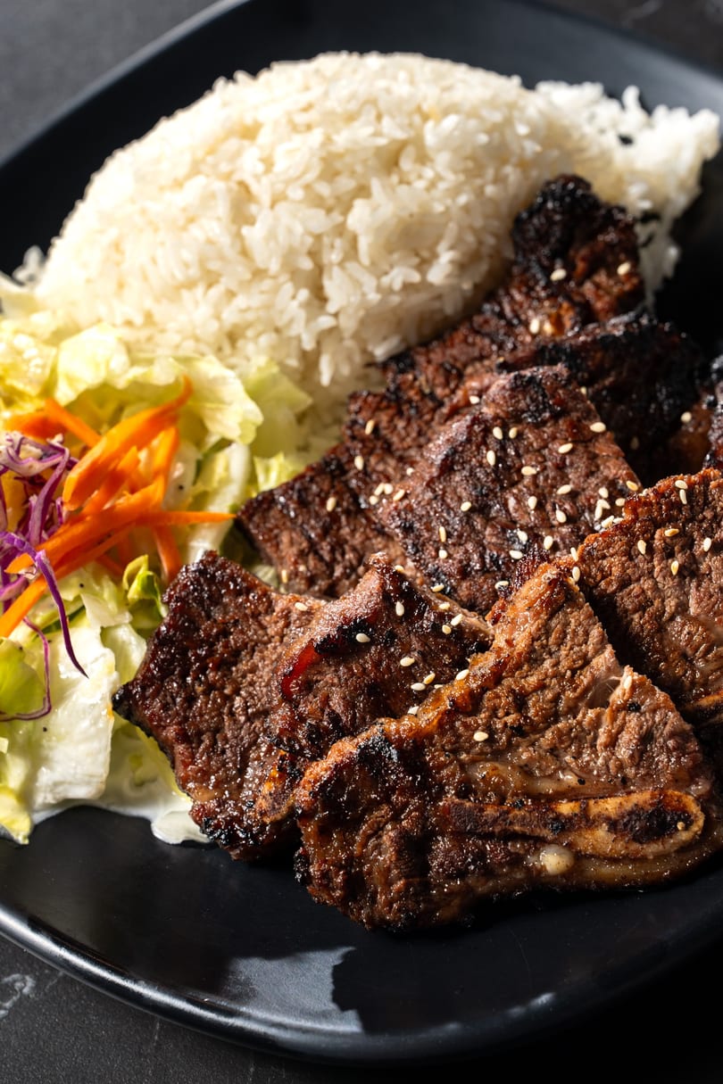 Grilled short ribs with sesame seeds, white rice, and a side salad on a black plate.