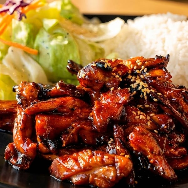 Teriyaki chicken with sesame seeds, white rice, and a side salad on a black plate.