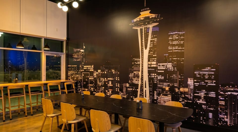 Restaurant interior with a long table, bar seating, and a large mural of the Seattle skyline.